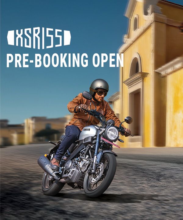 A helmeted rider in a brown jacket leans into a turn on a Yamaha XSR155 motorcycle beside a historic building.